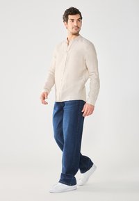 Chemise en lin beige clair à manches longues avec un col mandarin, devant boutonné ; associée à un jean en denim bleu foncé et des baskets blanches.