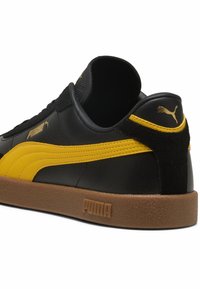 Fekete bőr sportcipők sárga kiegészítőkkel, texturált velúr sarkal, gumitalppal és dombornyomott Puma logóval a hátsó részen.