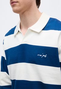 Άνδρας που φορά μπλε και άσπρη ριγωτή ριγέ ρουά μπλούζα με ανοιχτό γιακά και μικρό λευκό κεντητό κείμενο στο στήθος.