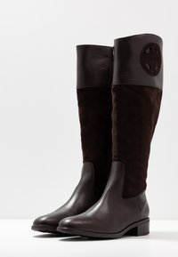 Stivali al ginocchio in pelle nera e suede marrone scuro. Presentano una finitura liscia, punta arrotondata, zip laterale e un emblema sulla parte superiore dello stivale.