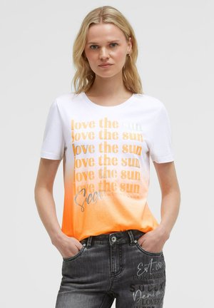 Woman wearing a white and orange ombre shirt with repeated text "love the sun" and dark gray jeans with printed text designs. Hands in pockets.