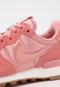 Tenisky Nike v jemné růžové z semiše a saténu, s hladkou gumovou podrážkou, odpovídajícími akcenty a výrazným logem na boku.