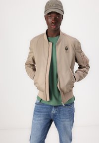 Beige bomber dzseki bordázott mandzsettákkal és hemmel, alatta zöld pólóval és kék farmernadrággal. A sapkán hímzett szöveg és kopott részletek találhatók.