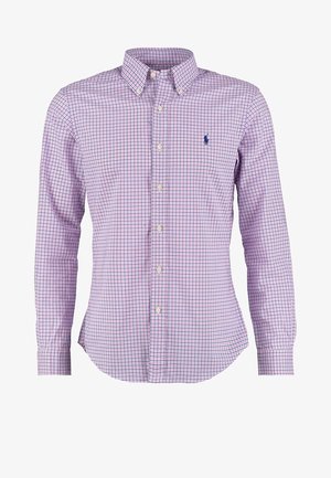 Mann steht mit Händen in den Taschen, trägt ein violett kariertes Hemd mit Knöpfen, beige Hosen und hellfarbige Turnschuhe.