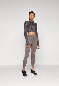 Crop top s dlouhým rukávem a tmavým vzorem v kombinaci s přiléhavými světle šedými legínami, obojí z elastické látky. Outfit doplňují černé sportovní boty.