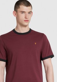 T-shirt bordeaux con colletto e polsini a costina neri. Presenta un piccolo logo giallo sulla parte sinistra del petto. Tessuto morbido, vestibilità standard.