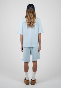 Personne aux longs cheveux ondulés portant une casquette noire, une chemise oversize bleu clair et un short, des chaussettes blanches et des chaussures marron, debout de dos.