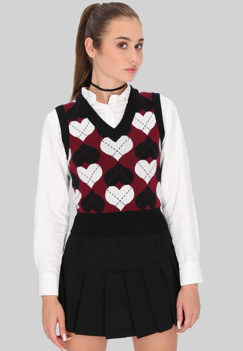 Pulover fără mâneci cu decolteu în V, cu un model de inimi roșii, negre și albe; purtat deasupra unei cămăși albe cu guler; asortat cu o fustă plisată neagră.