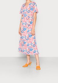 Blumiges Midikleid in Pink und Blau mit V-Ausschnitt, kurzen Schmetterlingsärmeln, Taillengürtel und schwingendem Rock. Kombiniert mit leuchtend orangefarbenen Absätzen.