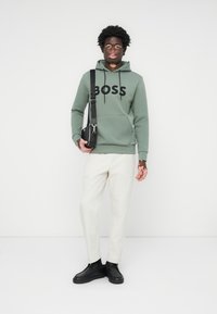 Sudadera verde con logo "BOSS" negro, bolsillo frontal, combinada con pantalones blancos y zapatillas negras. El modelo sostiene un pequeño bolso cruzado negro.
