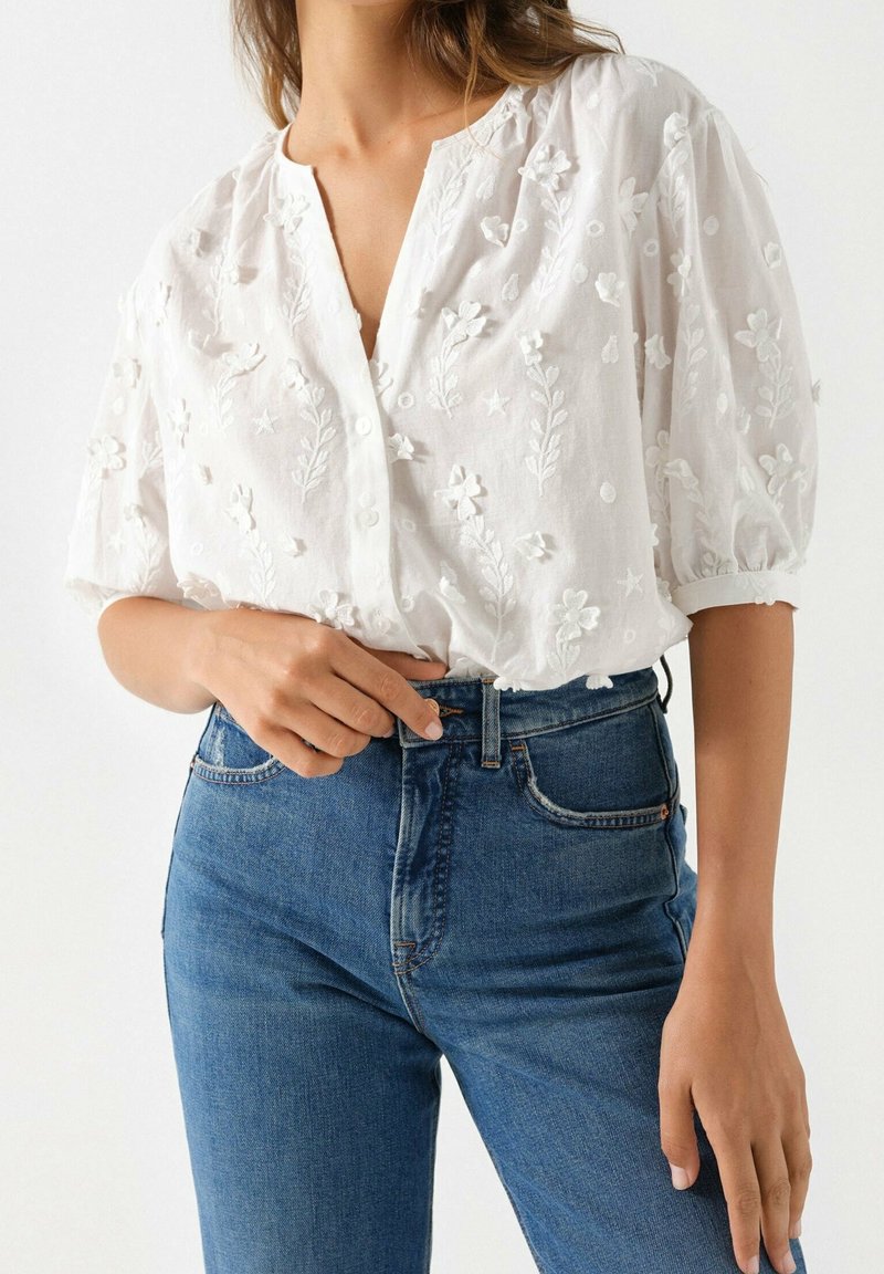 Hvid broderet bluse med blomster mønstre, V-udskæring, korte puffærmer, parret med høj midje blå jeans, der viser en klassisk denim tekstur.
