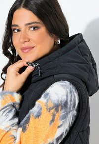 Svart vadderad väst med huva, med en dragsko i svart och vitt prickigt mönster. Modellen bär en färgglad, mönstrad tröja under.