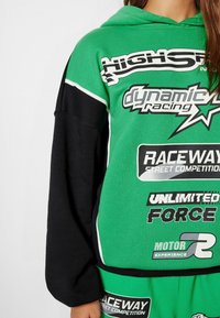 Grüner und schwarzer sweatshirt mit Kapuze, ausgestattet mit auffälligem weißen und schwarzen Text sowie rennsportinspirierten Grafiken. Teil eines passenden Sets.