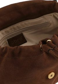 Innenraum einer braunen Veloursledertasche mit Kordelzugverschluss, ausgestattet mit einem Reißverschlussfach, beigem Futter und einem kleinen Etikett mit der Aufschrift „abro.“