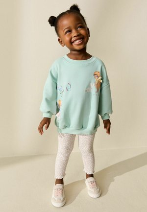 Uśmiechnięte małe dziecko w jasnoniebieskiej bluzie z nadrukami z kreskówek, legginsach w wzory i różowych tenisówkach, stojące na tle jednolitego beżowego tła.