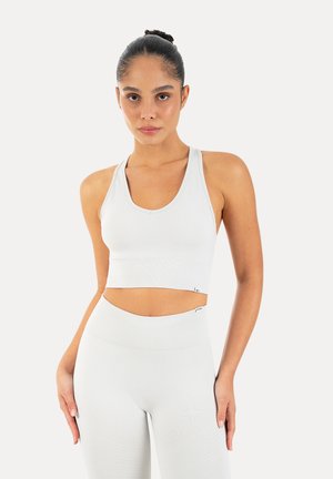 Kobieta z włosami upiętymi w kok, ubrana w biały bezrękawnik typu crop top i legginsy z wysokim stanem, stojąca na tle jednolitego jasnego tła.