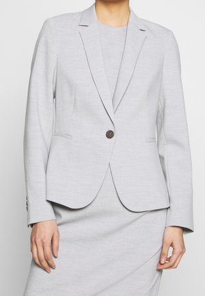 Woman wearing a light grey tailored blazer with a single dark button, matching skirt, and a round-neck top underneath.