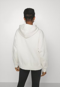 Felpa bianca con finitura morbida e testurizzata. Presenta un grande cappuccio e una vestibilità rilassata, abbinata a leggings neri. Design semplice e privo di decorazioni.