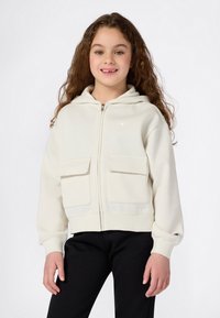 Ragazza sorridente con lunghi capelli ricci, che indossa una felpa cremosa con zip e tasche frontali e pantaloni neri, in piedi su uno sfondo bianco.