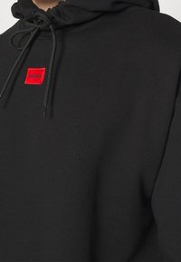 Schwarzer Hoodie aus weichem Stoff mit einem roten quadratischen Aufnäher mit der Aufschrift "HUGO" auf der linken Seite. Verfügt über eine Kapuze mit Kordelzug und einen lockeren Sitz.