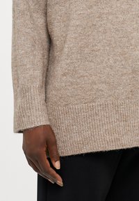 Maglione beige a maglia con orlo e polsini a coste, con una finitura morbida e testurizzata e una vestibilità comoda. Mano mostrata per scala.