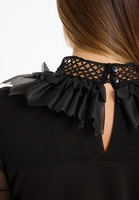 Schwarzes Kleidungsstück mit einem strukturierten Netzkragen, der mit einem fransigen schwarzen Akzent und zwei schwarzen Knöpfen im Rücken versehen ist.