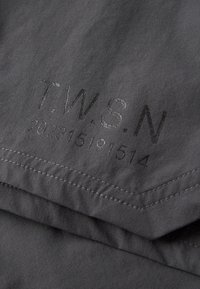 Dunkelgrauer Stoff mit genähtem Saum, auf dem in schwarzem Drucktext "T.W.S.N" und eine Zahlenfolge "202315191514" abgebildet sind.