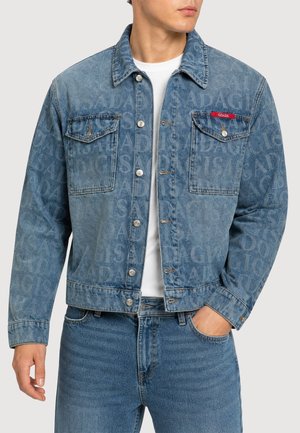 Mann trägt blaue Jeansjacke und Jeans, mit dem Textmuster "GISADA" auf der Jacke und einem roten Etikett auf der linken Brusttasche.