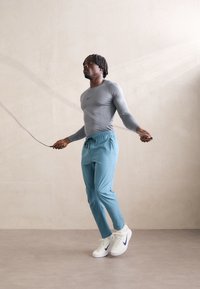 Conjunto deportivo que incluye una camiseta de manga larga gris, pantalones de chándal azul claro y zapatillas Nike blancas. Modelo saltando la cuerda en un ambiente interior.