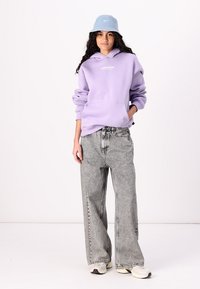 Lilla hettegenser med frontlogo, kombinert med lysegrå vide jeans og en lyseblå bøttehatt. Skoene er kremfargede med mørke detaljer.
