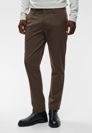 Homme portant un pantalon marron sur mesure, des bottines Chelsea en cuir noir et un haut gris clair à manches longues, debout sur fond blanc.