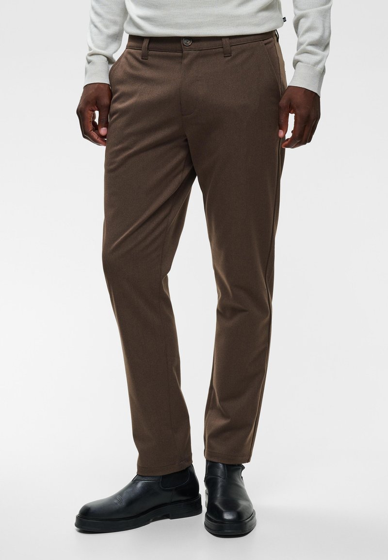 Homme portant un pantalon marron sur mesure, des bottines Chelsea en cuir noir et un haut gris clair à manches longues, debout sur fond blanc.