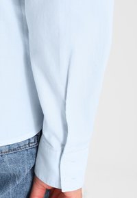 Pale blue long-sleeve shirt with a textured fabric, featuring buttoned cuffs and a straight hem, paired with denim jeans.