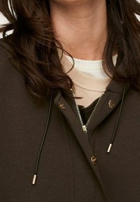 Veste marron foncé à fermeture éclair avec une texture douce, dotée d'accents dorés, de cordons réglables et d'une doublure intérieure crème en couches.