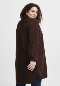 Abrigo de forro polar marrón con un alto cuello, mangas largas y un bolsillo lateral con cremallera. Textura suave y silueta alargada con dobladillo curvado.