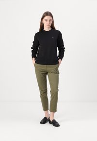 Svart hettegenser med rund hals, paret med olivengrønne chinos. Løs passform. Modellen har på seg svarte slip-on sko.