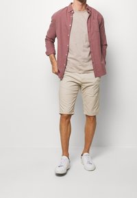 Mężczyzna w zakurzonym różowym koszuli z guzikami, beżowej koszulce, beżowych szortach i białych trampkach stoi pod białą ścianą.