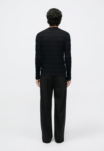 Μαύρο πλεκτό πουλόβερ με ριγέ υφή και μακριά μανίκια, φορεμένο με μαύρο παντελόνι με ψηλή μέση. Ορατό υφαντό μοτίβο στην επιφάνεια του πουλόβερ.