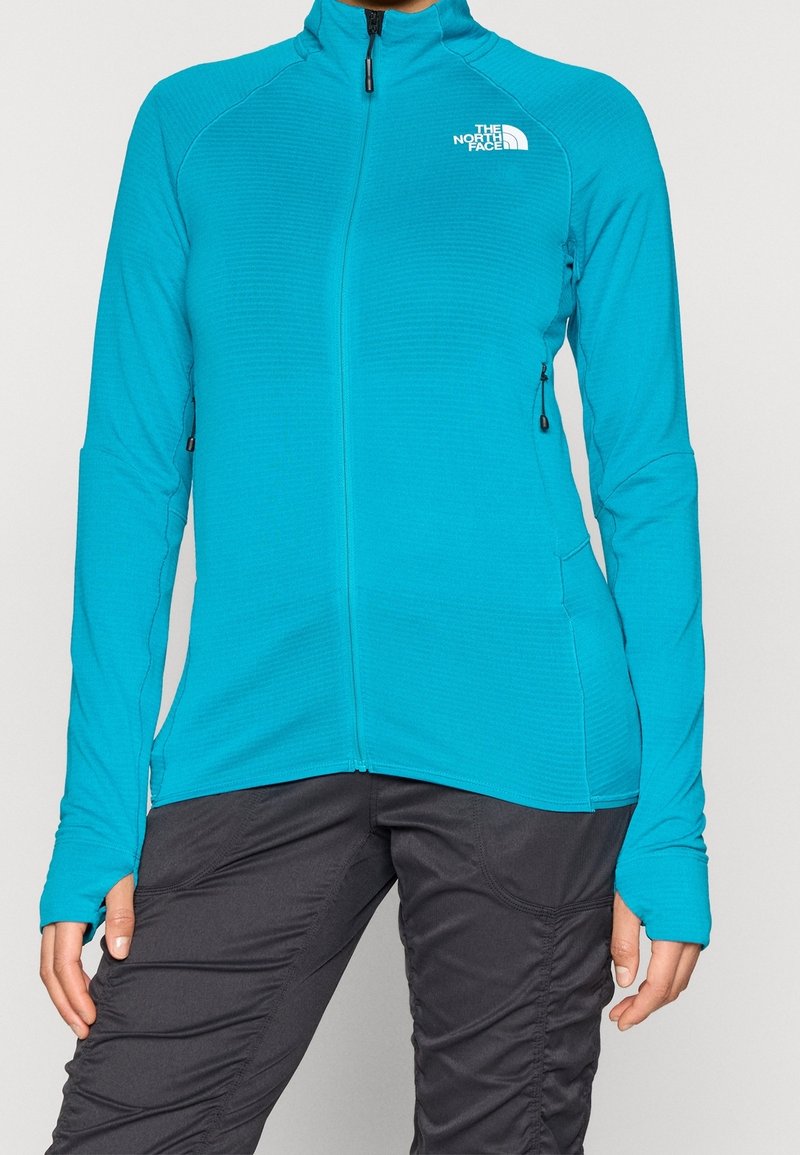 Gros plan d'une personne portant une veste zippée bleu vif avec des trous pour les pouces et un pantalon noir, montrant le logo The North Face sur la poitrine.