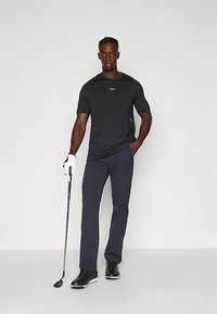 T-shirt preta de manga curta com logótipo, calças de golfe azul-marinho, luva de golfe na mão esquerda, segurando um taco de golfe, e sapatos desportivos pretos.