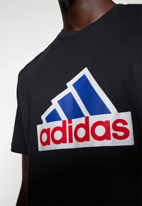 Černé tričko s krátkými rukávy, s velkým logem Adidas v bílé, modré a červené barvě. Materiál vypadá hladce a má přiléhavý střih.