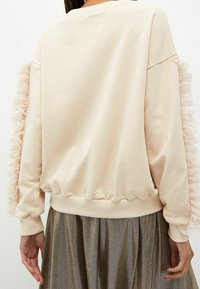 Sudadera beige de manga larga con detalles de tul fruncido a lo largo de las mangas, combinada con una falda plisada metálica.