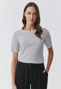 Witte top met korte mouwen en zwarte horizontale strepen, ronde halslijn. Gecombineerd met hoge zwarte broeken met een knoopdetail.