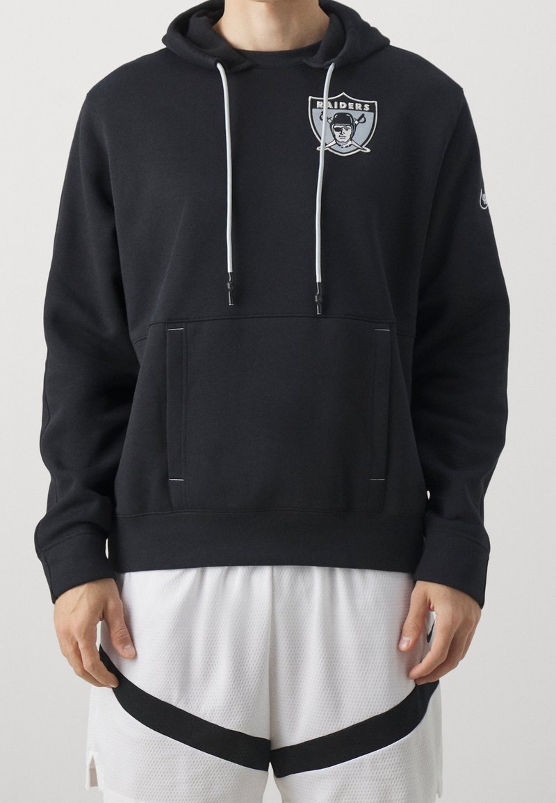 Personne portant un sweat à capuche Raiders noir avec des cordons blancs et un short de basket blanc avec une bande diagonale noire.
