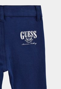 Pantalons en tissu bleu marine avec un logo "GUESS" brodé blanc, ornés de détails décoratifs et d'une texture lisse.