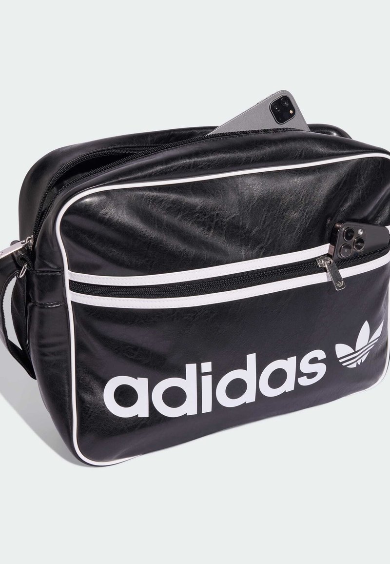 Black faux leather bag with white accents, zippered pocket, and bold "adidas" logo. Features a structured rectangular shape and adjustable strap.