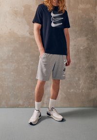 Person klädd i svart Nike t-shirt, grå Nike shorts, vit Nike sneakers och vita ankelsockor som står mot en texturerad vägg.