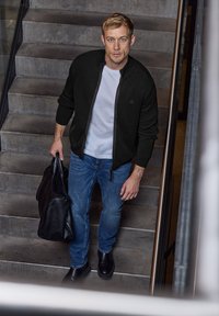 Zippad svart bomberjacka, vit t-shirt, blå jeans och svarta stövlar. Håller en stor svart väska. Står på betongtrappor med ett metallräcke.