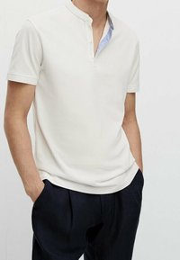 Vit kortärmad pikétröja med ståkrage, som har ett blått mönstrat foder vid halsringningen och en slät, strukturerad yta.