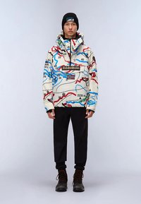 Chaqueta multicolor con diseño de mapa, que incluye capucha y cremallera, combinada con pantalones negros y botas marrones. Gorro negro en la cabeza.